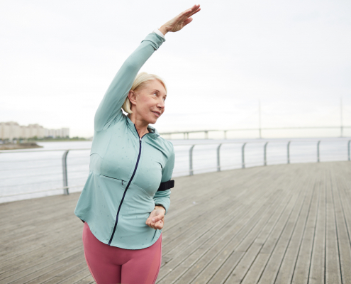 Mature active female in sportswear doing side-bends while training by riverside in urban environment on summer morning