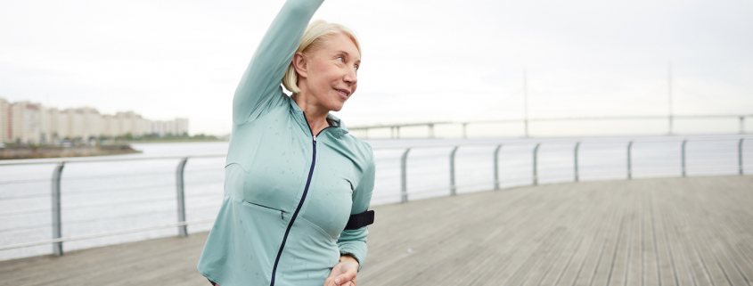 Mature active female in sportswear doing side-bends while training by riverside in urban environment on summer morning