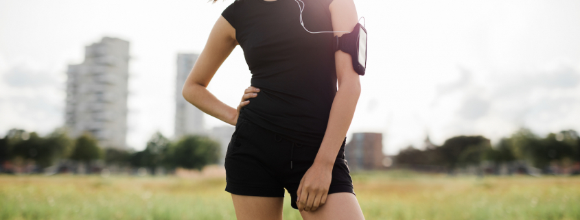Cropped shot of fitness woman standing in urban park wearing sportswear and mobile phone armband. Female model in sportswear with earphones listening to music during workout break.