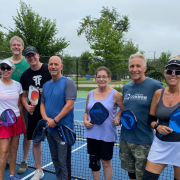 Group of people smiling and standing together in front of a pickleball net.