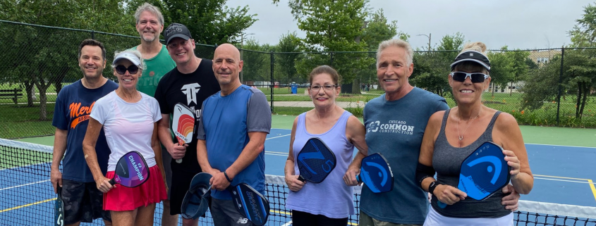 Group of people smiling and standing together in front of a pickleball net.